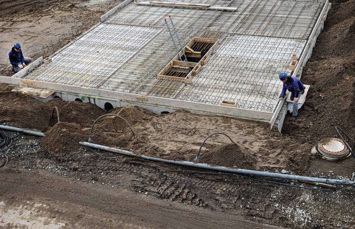 Eine Baustelle zeigt zwei Bauarbeiter, die an einer teilweise verlegten Betonfundamentplatte arbeiten. Erde und Baugeräte sind sichtbar.