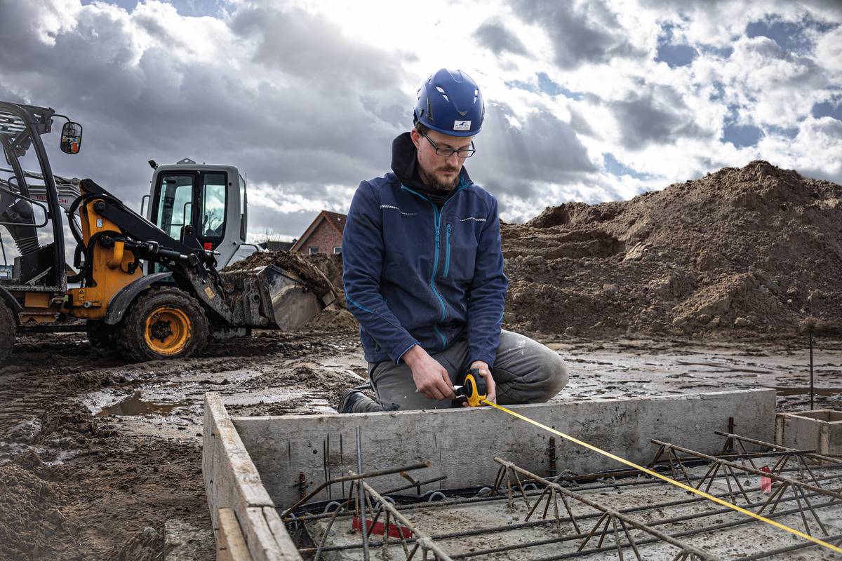 Mann mit Helm misst Betonfundament mit Maßband auf Baustelle; Bagger im Hintergrund bei bewölktem Himmel.