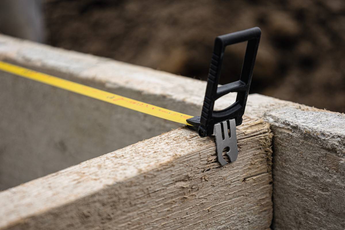 Ein Maßband wird über eine Holzplatte gezogen, um eine Länge zu messen. Der Hintergrund ist verschwommen und zeigt Baustellenmaterialien.