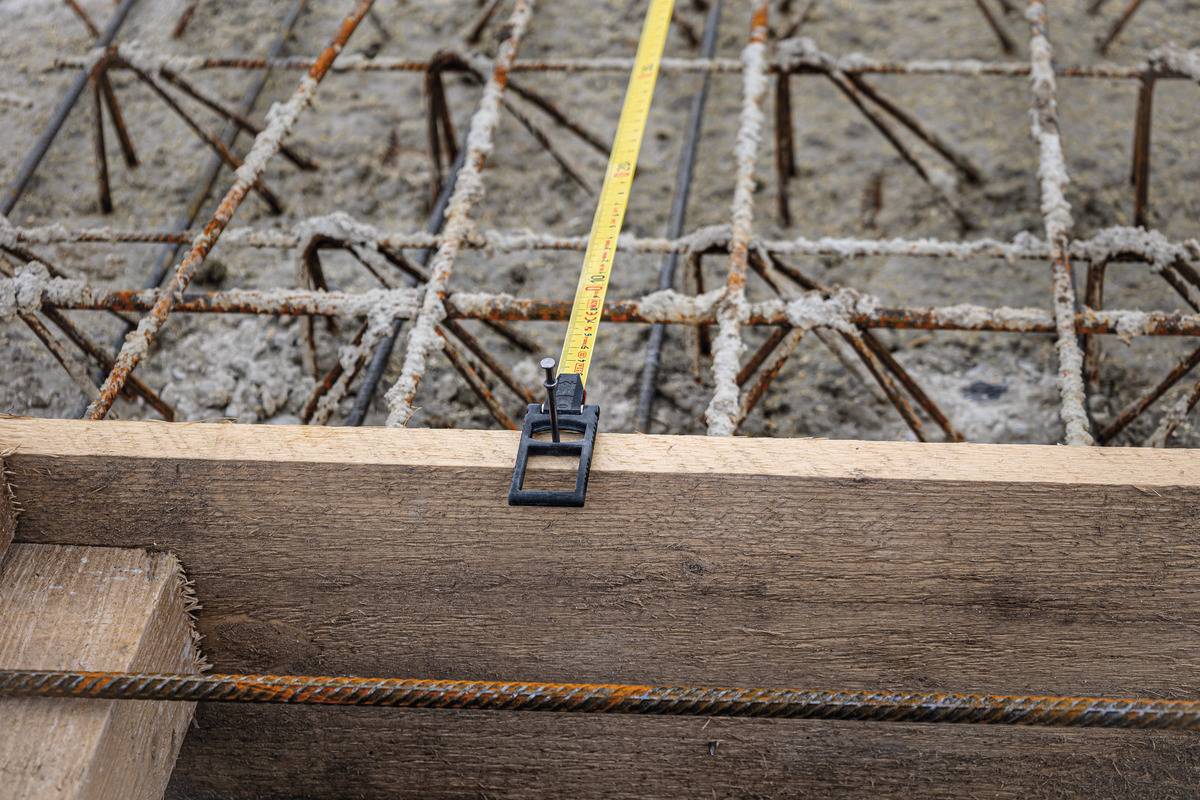 Ein gelbes Maßband liegt auf einer Baustelle über Stahlstreben und Beton, zeigt eine Länge im Aufbauprozess an.