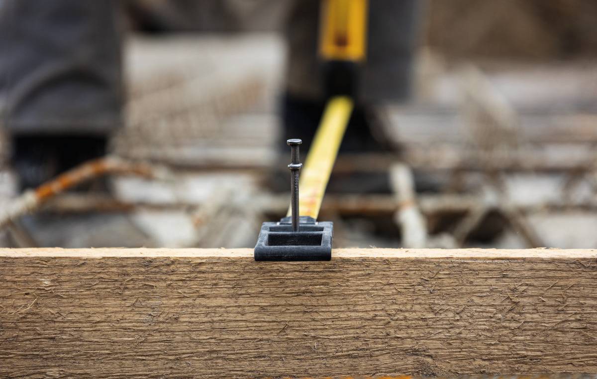 Holzbalken mit eingestecktem Nagel und aufgelegtem Maßband, Schärfe auf Nagelspitze, unscharfer Hintergrund mit Metallstreben.