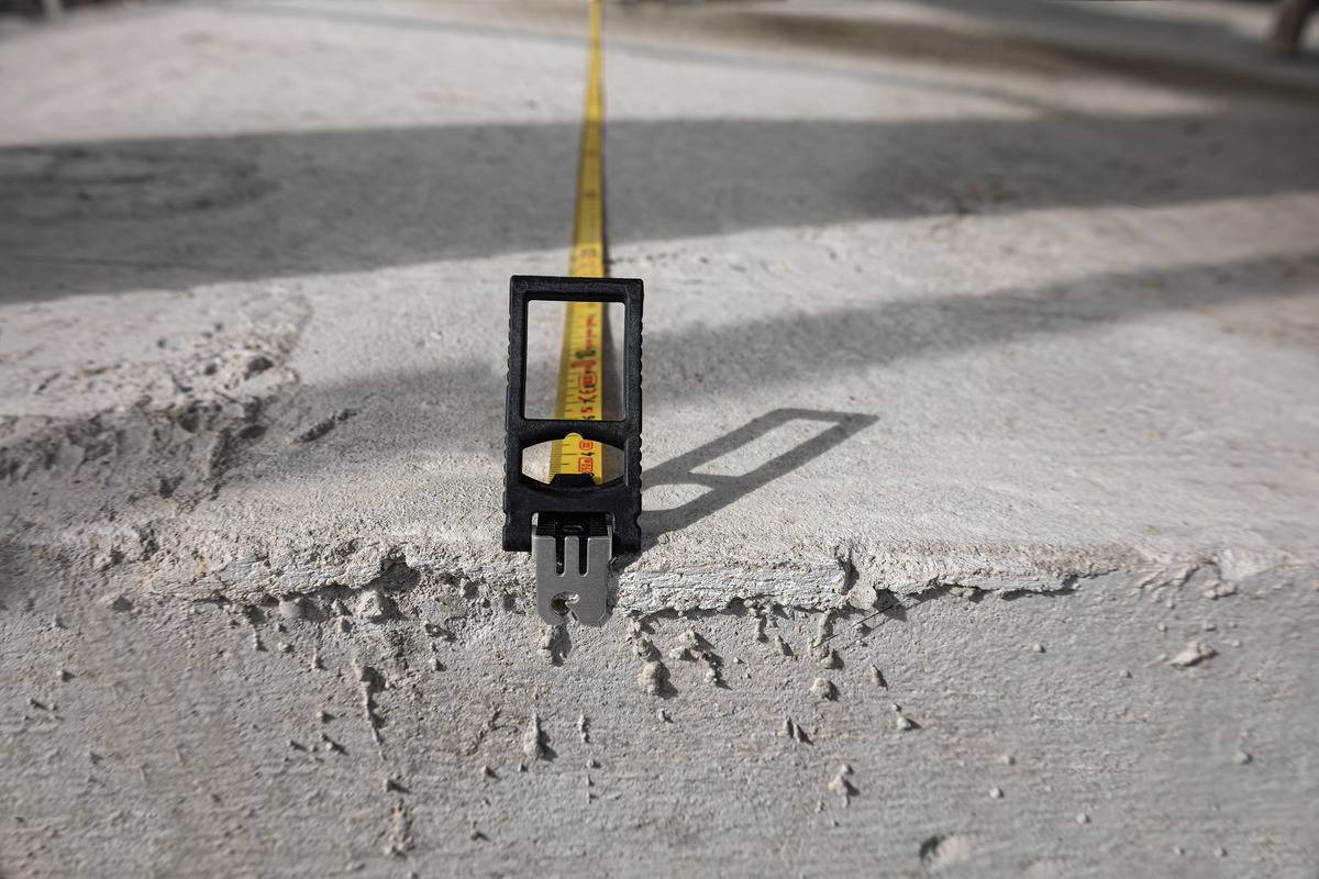 Ein Maßband wird auf Betonboden in die Ferne gezogen, um eine Länge zu messen. Die Oberfläche ist rau und uneben.