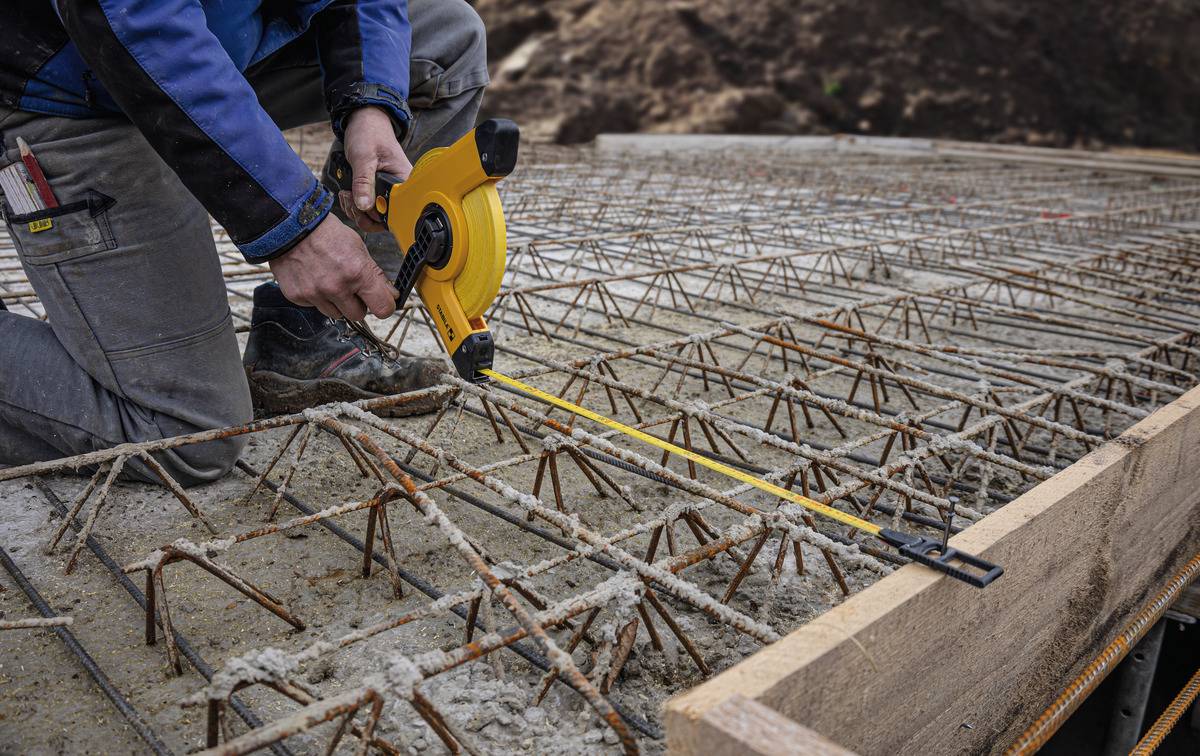 Eine Person misst mit einem Maßband auf einer Baustelle die Bewehrung eines Betonfundaments. Die Konstruktion ist für Stabilität vorbereitet.