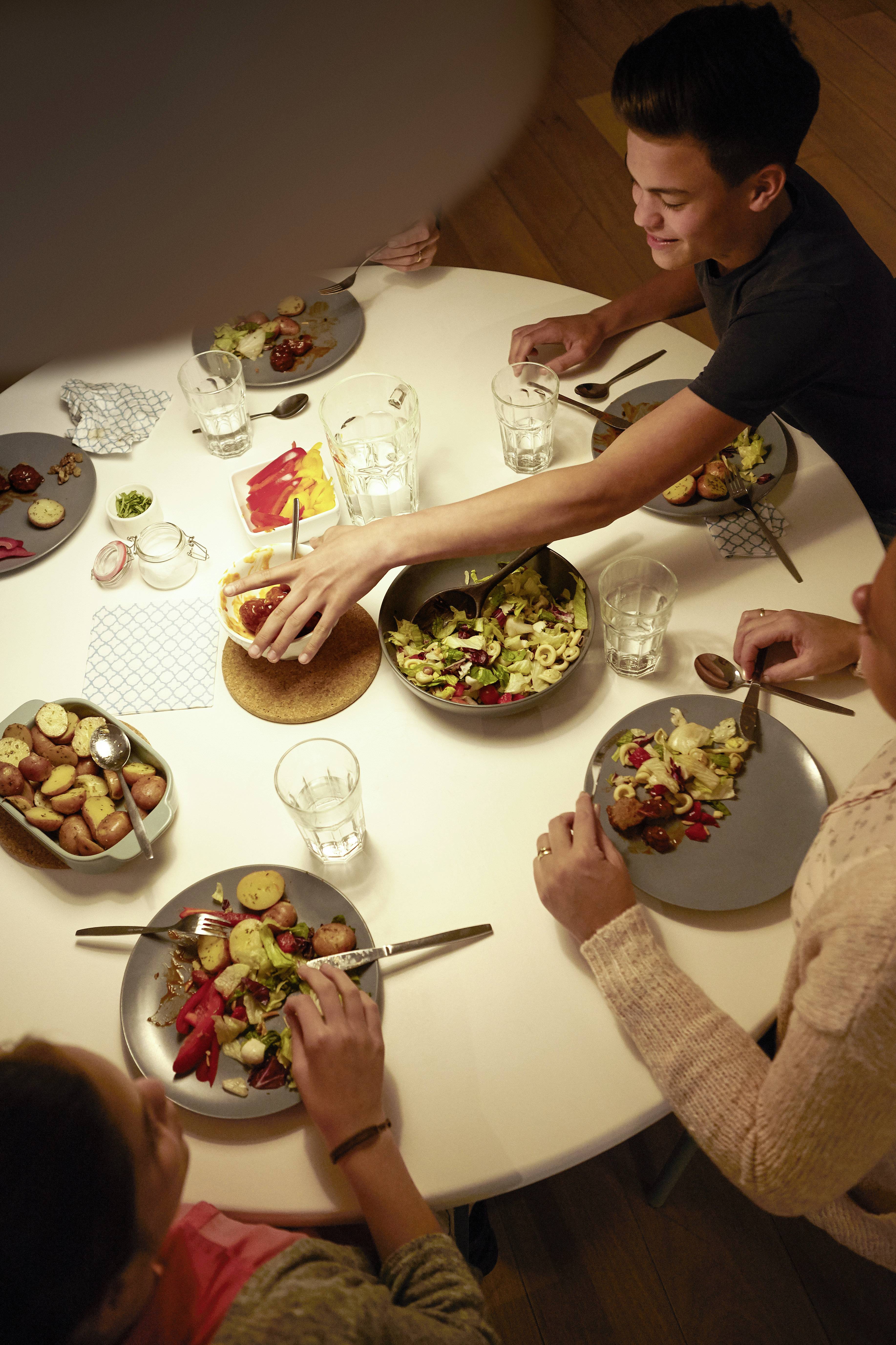 Eine Gruppe von Personen sitzt an einem runden Tisch und isst ein gemeinsames Essen. Auf dem Tisch stehen verschiedene Salate und Beilagen.