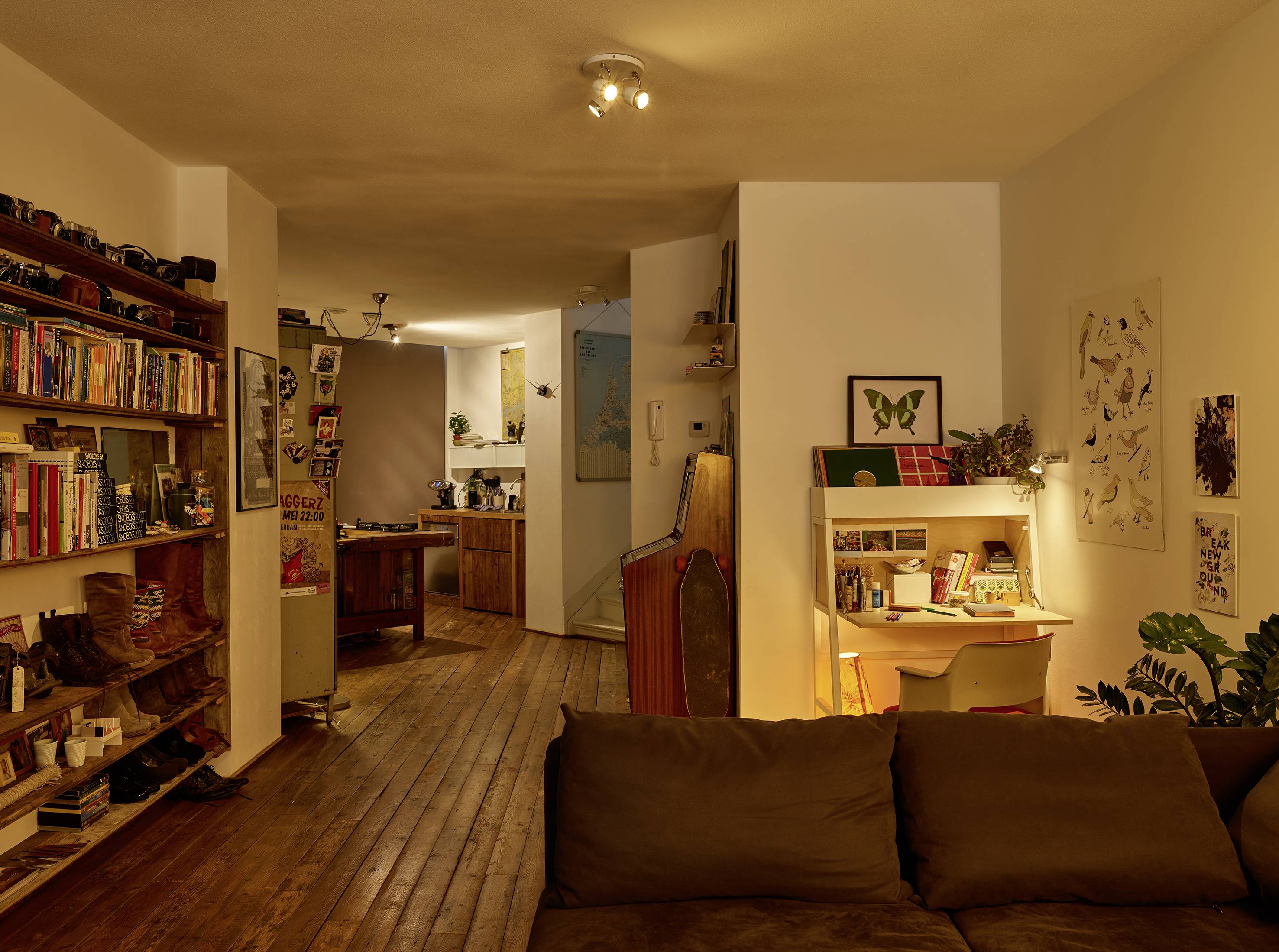 Gemütlich eingerichtetes Wohnzimmer mit Büchernregal, Schreibtisch und Sofa; warme Beleuchtung schafft eine einladende Atmosphäre.
