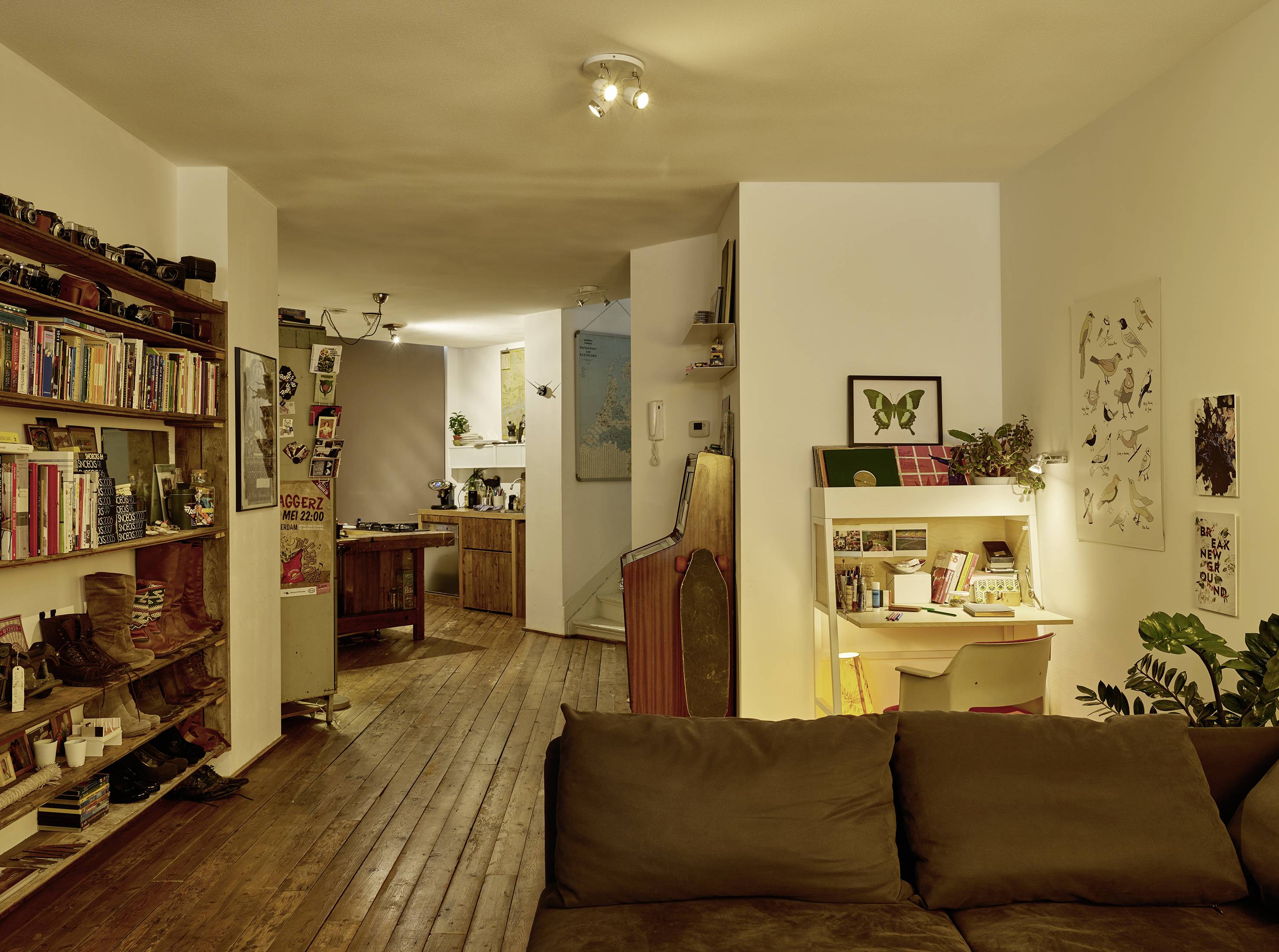 Ein gemütliches Wohnzimmer mit Holzboden, bestehend aus einem Sofa, Bücherregalen, einem kleinen Schreibtisch und Pflanzen. Im Hintergrund ist eine Küche sichtbar.