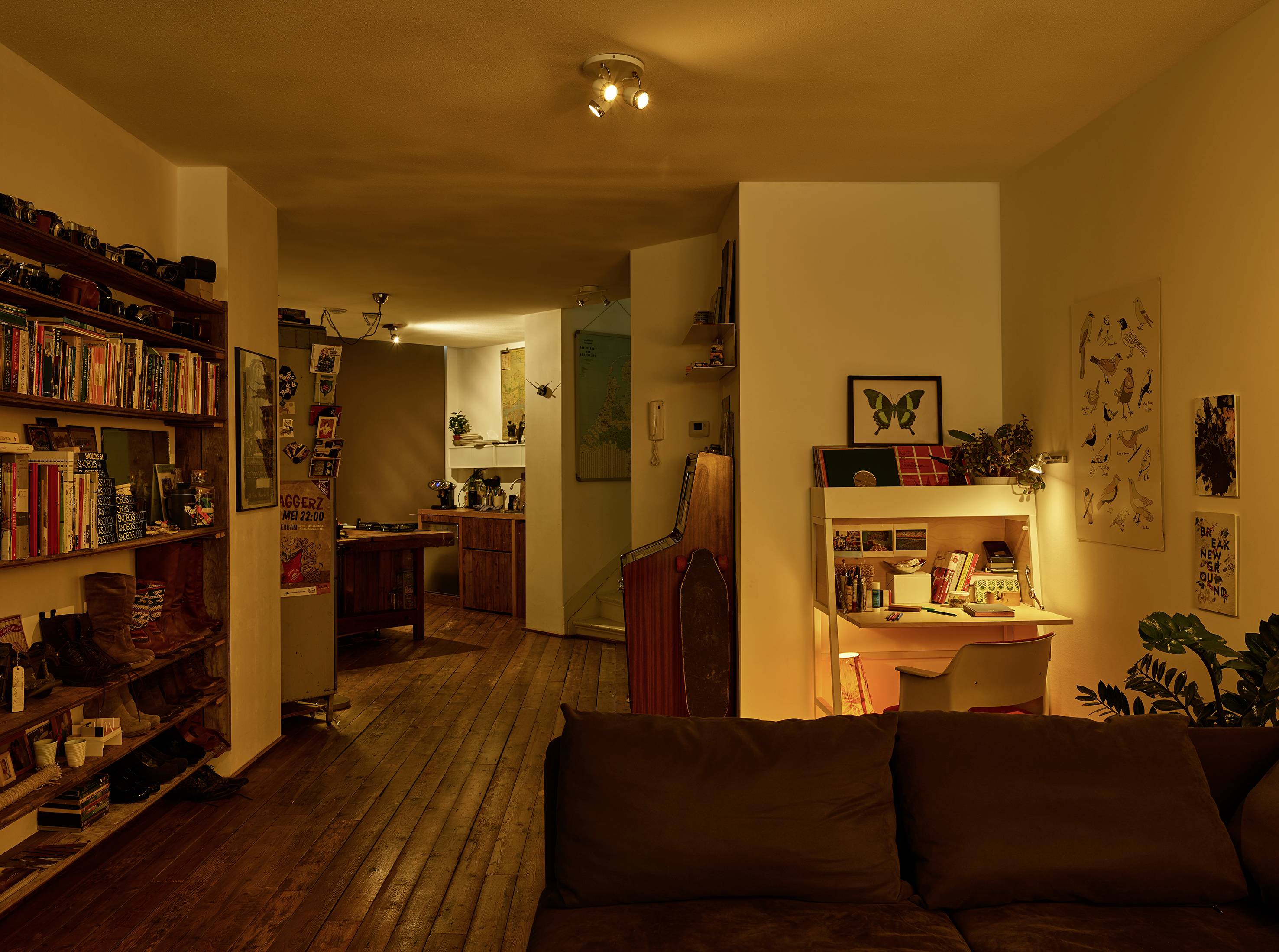 Gemütliches Wohnzimmer mit Holzboden, Regal mit Büchern und Pflanzen, Sofa und Schreibtisch mit Lampe in gedämpfter Beleuchtung.