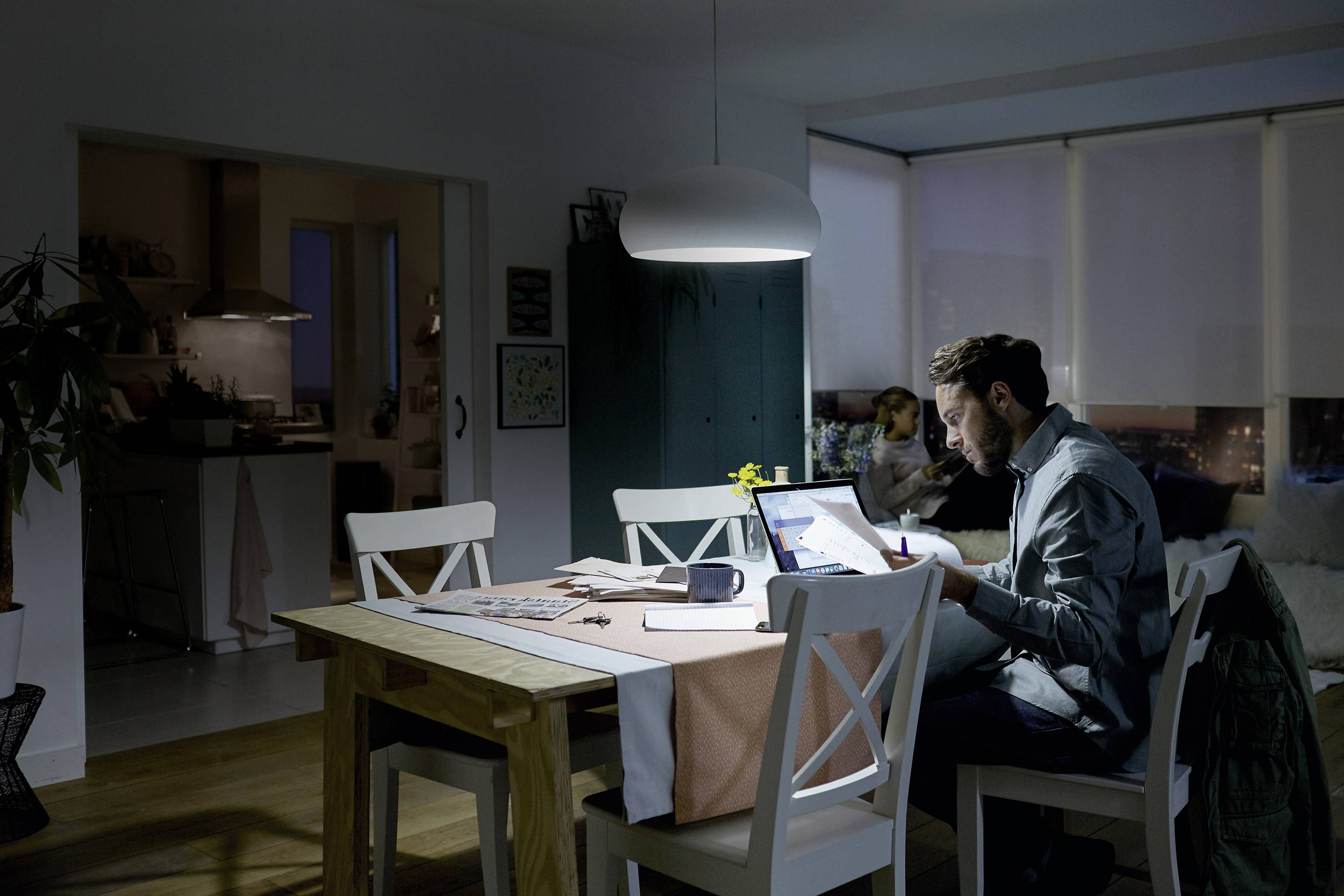 Ein Mann arbeitet abends konzentriert an einem Laptop am Esstisch, während im Hintergrund jemand auf dem Sofa entspannt sitzt.