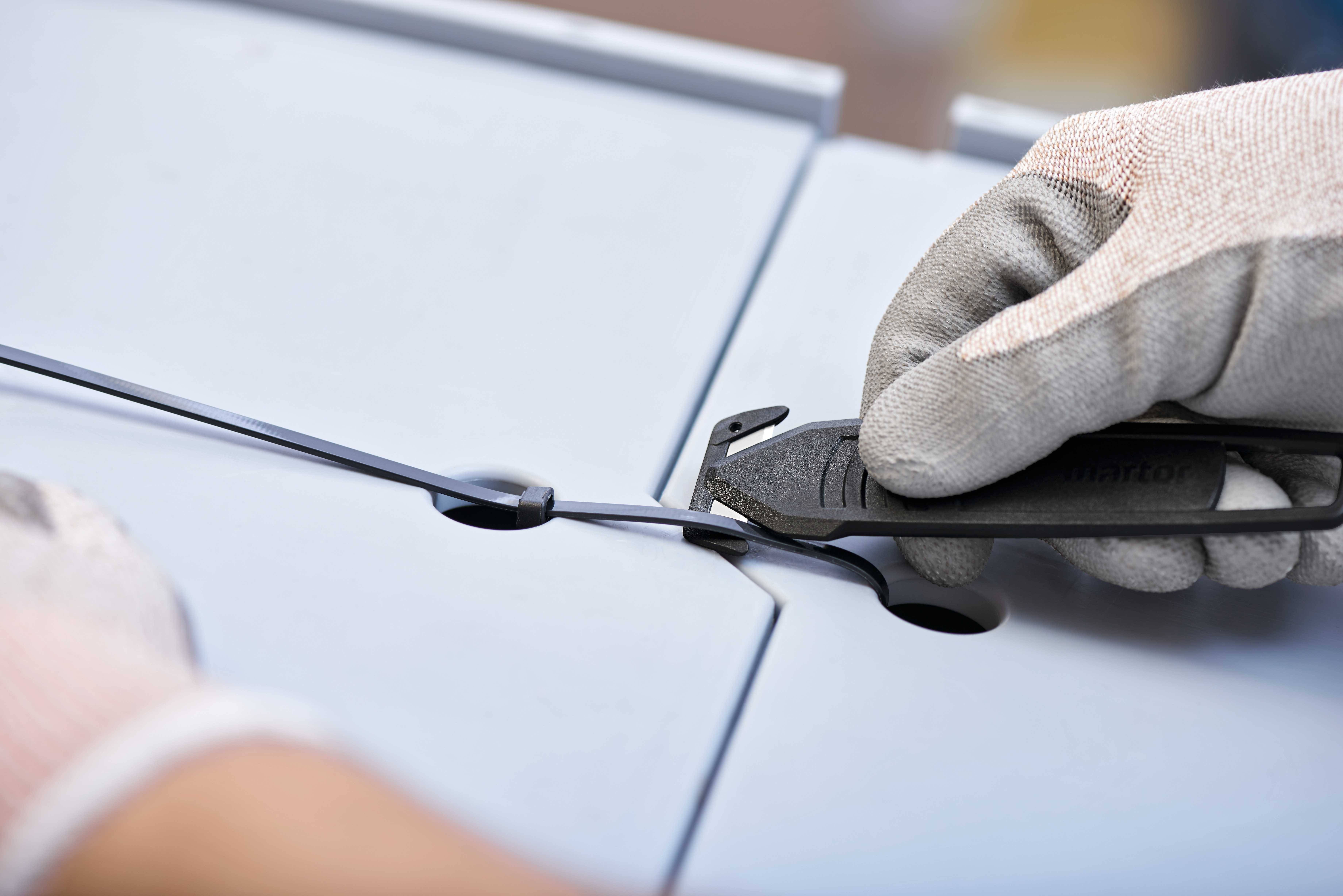 Eine Person in Handschuhen schneidet mit einem Messer einen Kabelbinder durch, der zwei graue Platten zusammenhält.