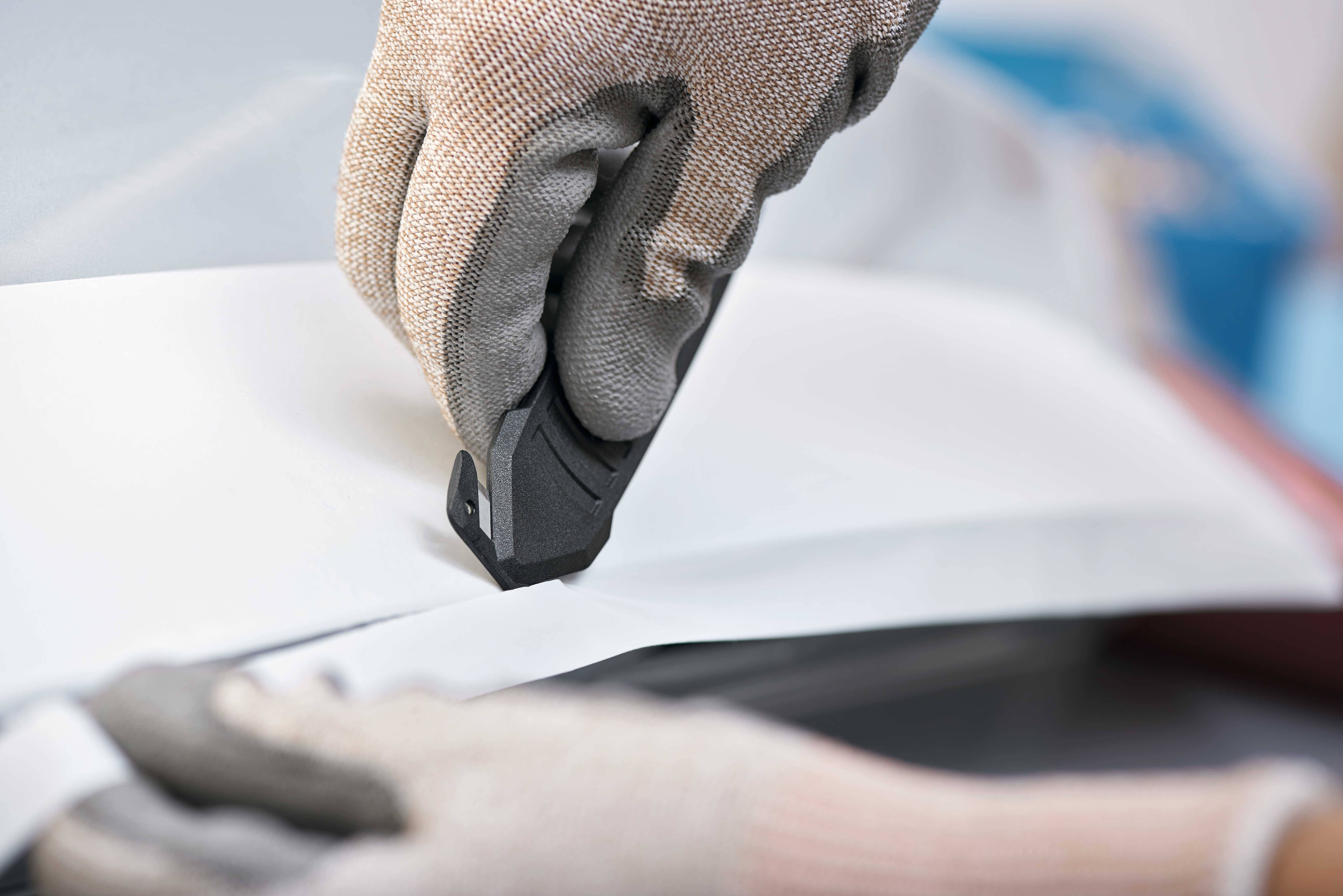 Eine Person mit Handschuhen schneidet mit einem Cuttermesser durch dickes Papier oder Karton auf einer glatten Oberfläche.