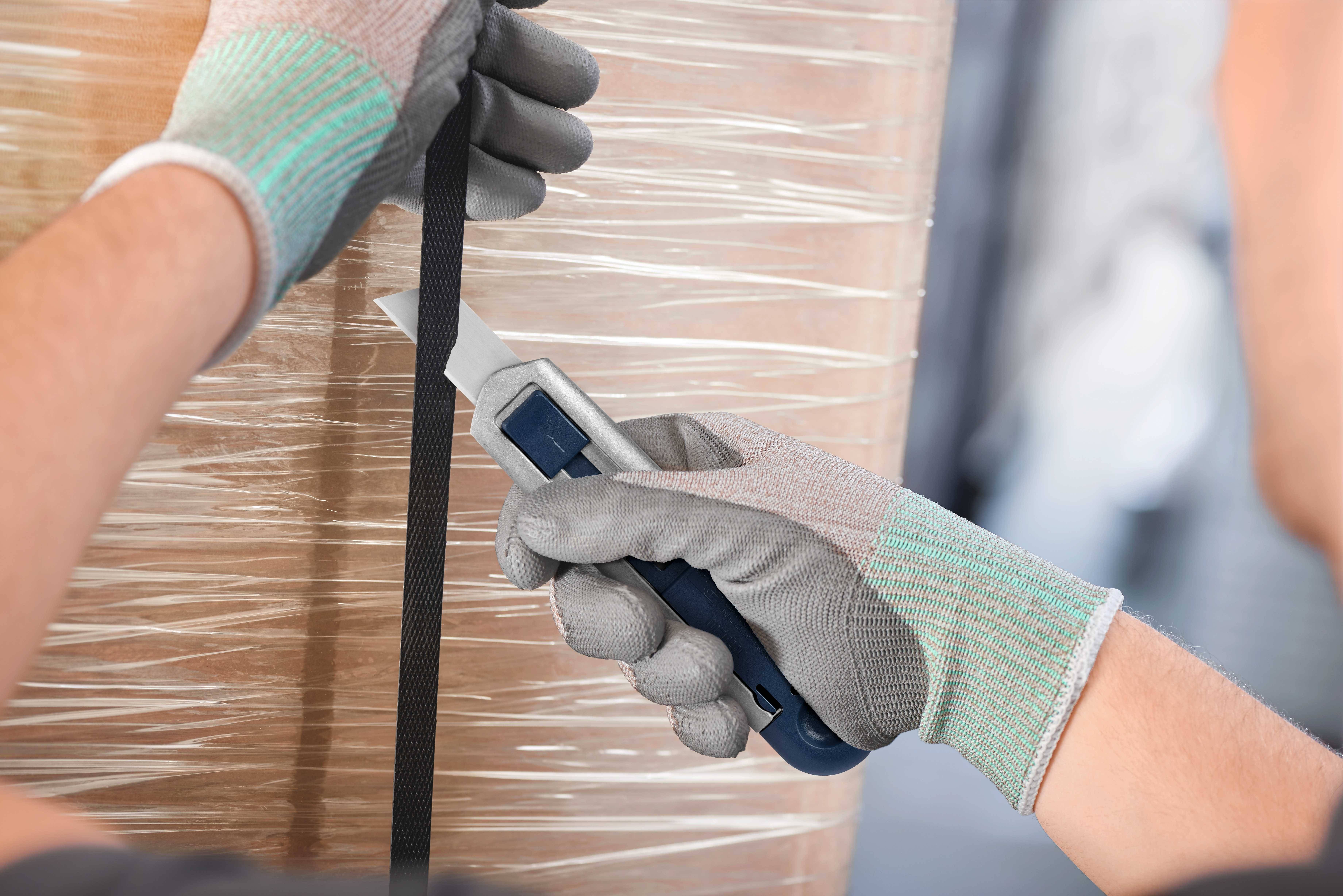 Nahaufnahme von Händen mit Schutzhandschuhen, die einen Sicherheitsmesser verwenden, um eine schwarze Plastikbandage auf einer Palette zu durchtrennen.