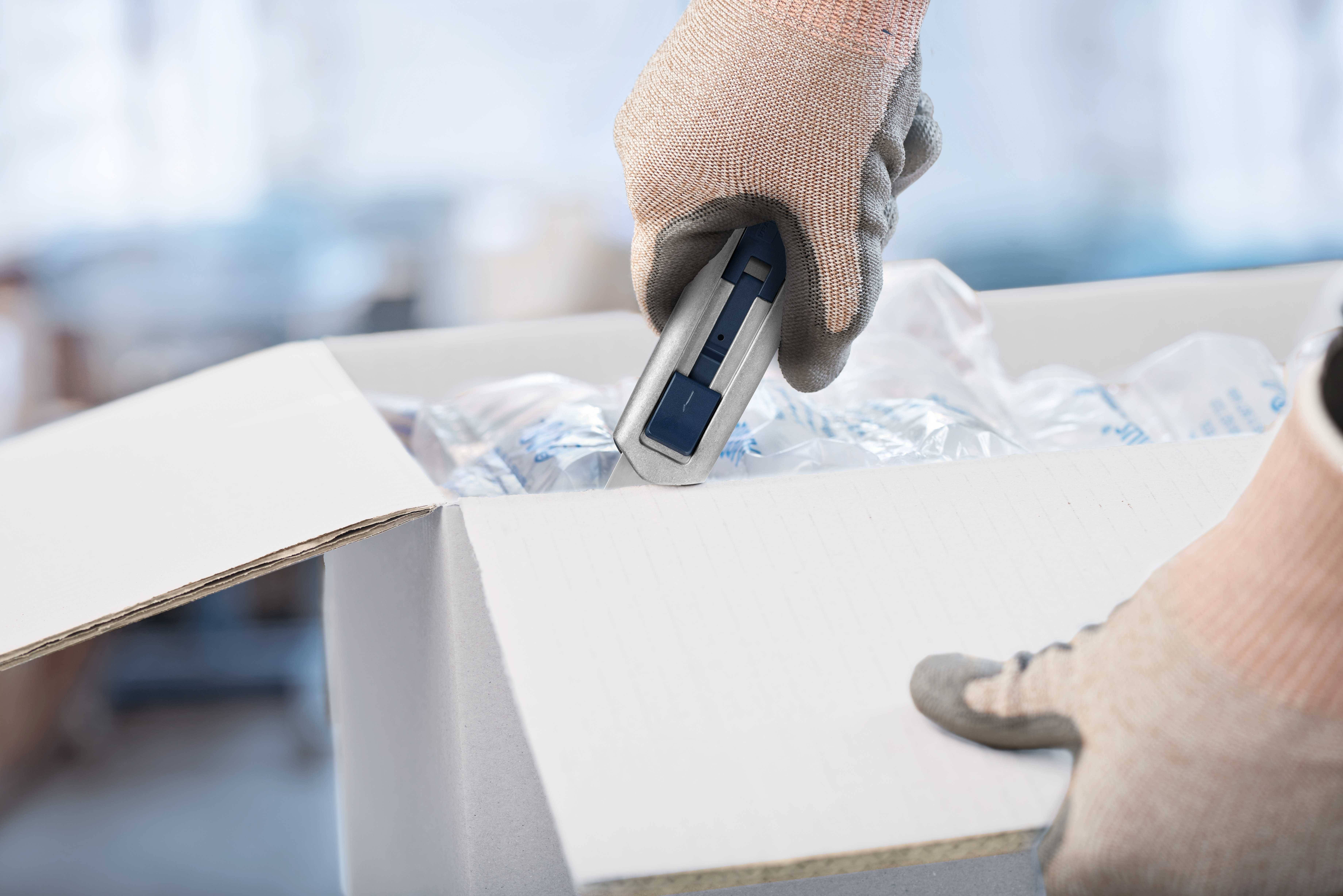 Eine Person mit Handschuhen öffnet einen Karton mit einem Cuttermesser. Innenmaterial zum Schutz des Inhalts ist sichtbar.