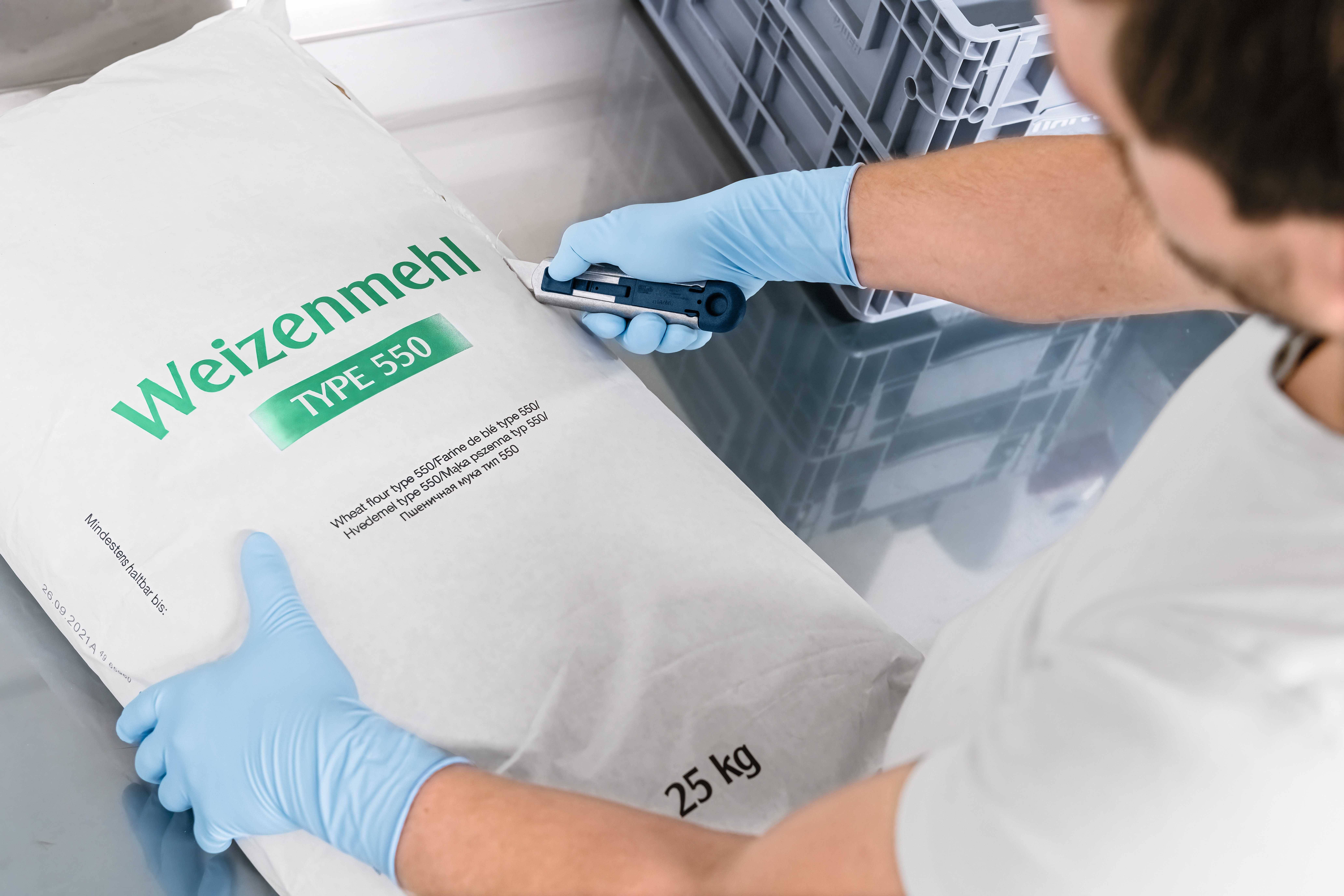 Ein Mann mit blauen Handschuhen öffnet einen 25-kg-Sack Weizenmehl Type 550 mit einem Cutter-Messer.