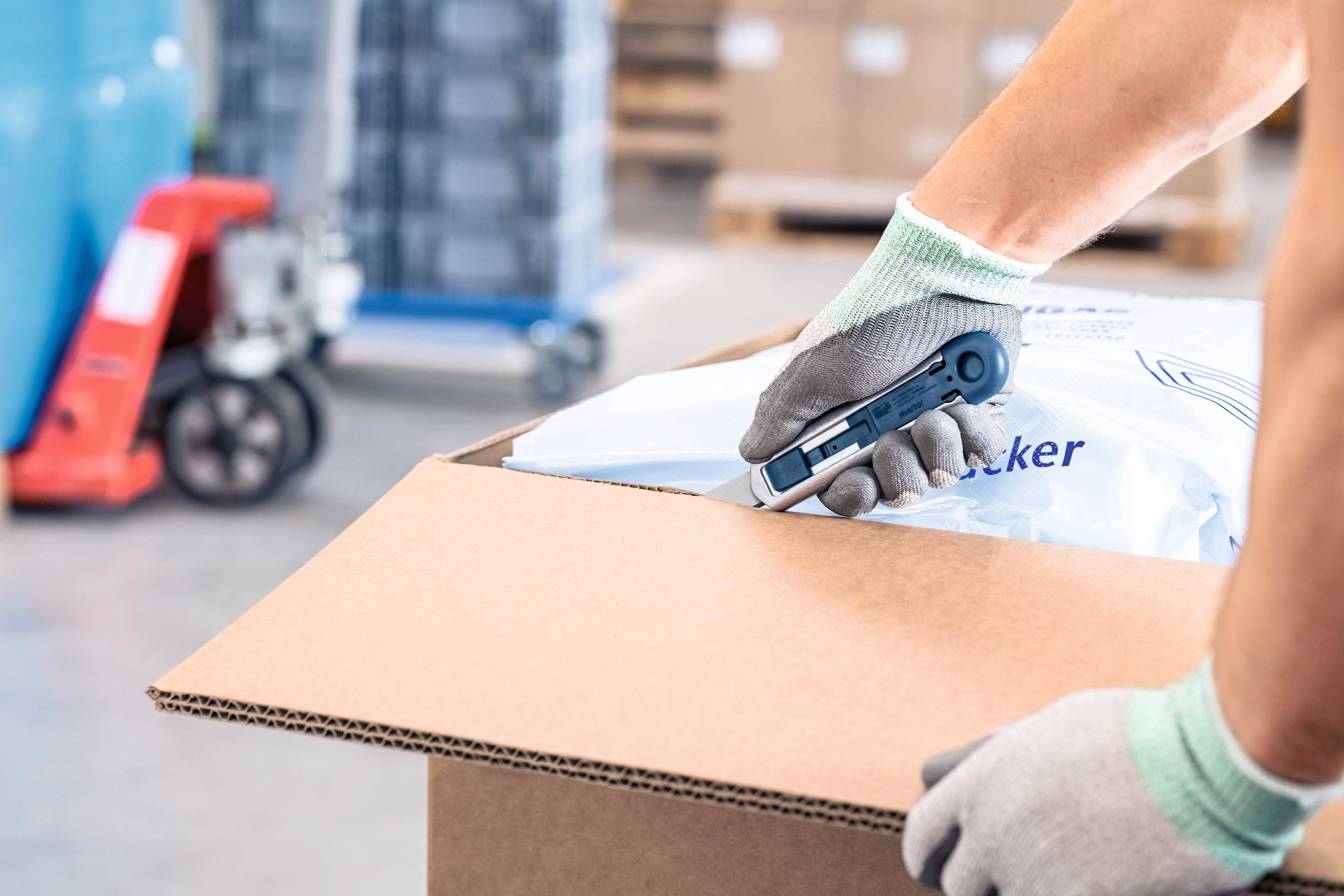 Eine Person öffnet mit einem Cuttermesser ein Kartonpaket. Die Szene findet in einer Lagereinrichtung statt.