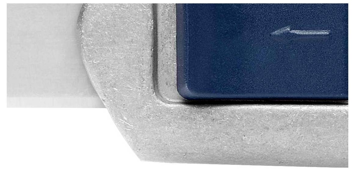 Nahaufnahme einer Metallkante neben einer dunklen Fläche mit einem kleinen eingravierten Pfeil, der nach links zeigt.