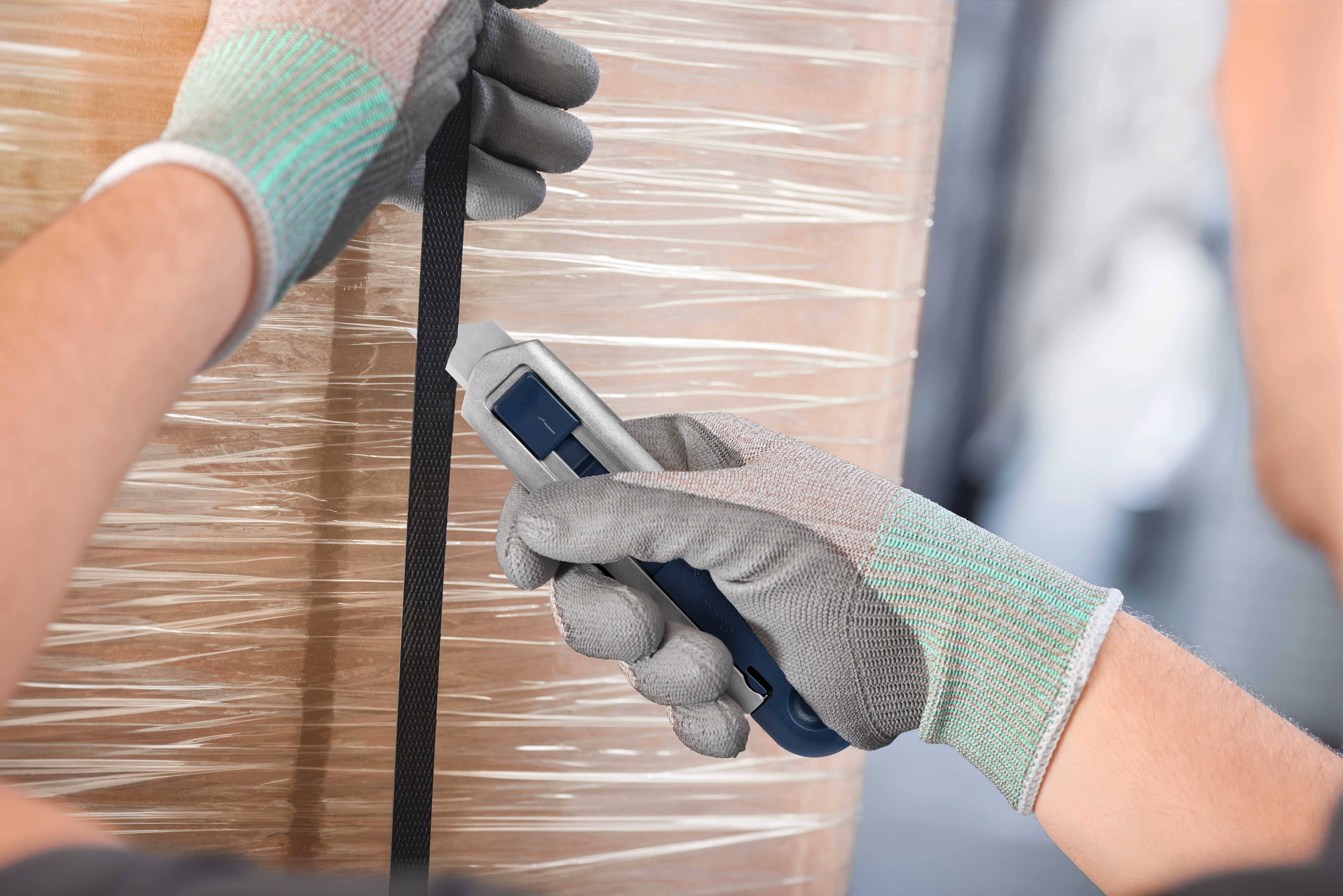 Eine Person in Handschuhen schneidet mit einem Messer ein schwarzes Band an einer mit Folie umwickelten Palette.