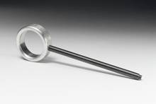 Ein Metallring ist mit einem langen, dünnen Stab verbunden. Das Ensemble liegt auf einer hellen Oberfläche mit dunklem Hintergrund.