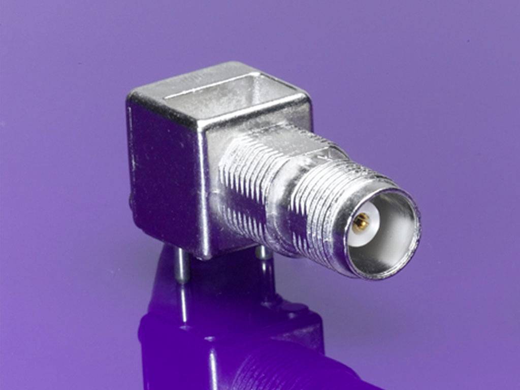 Ein silberner Koaxial-Stecker mit quadratischem Gehäuse auf lila Hintergrund, geeignet für elektronische Verbindungen.