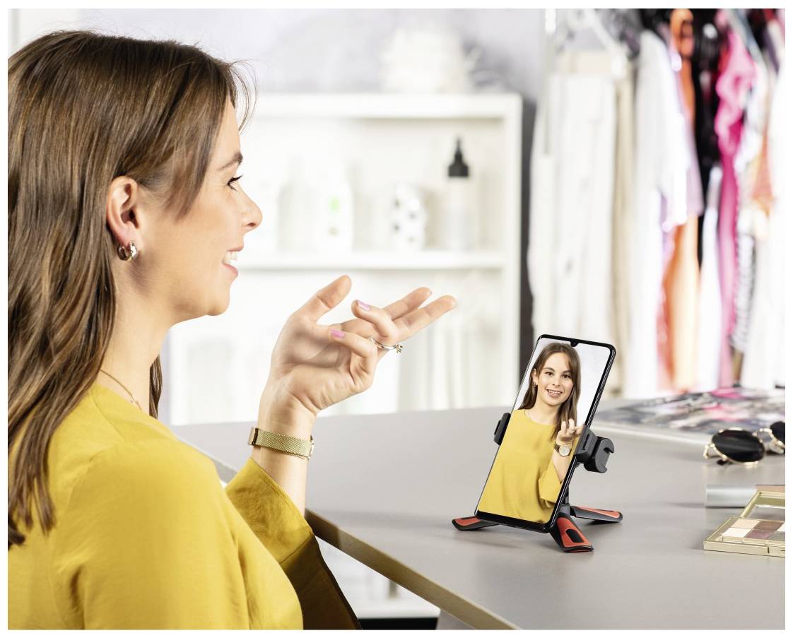 Frau in gelber Bluse videochattet auf Smartphone, das auf einem Tisch steht. Kleidungsstücke und weiße Regal im Hintergrund.
