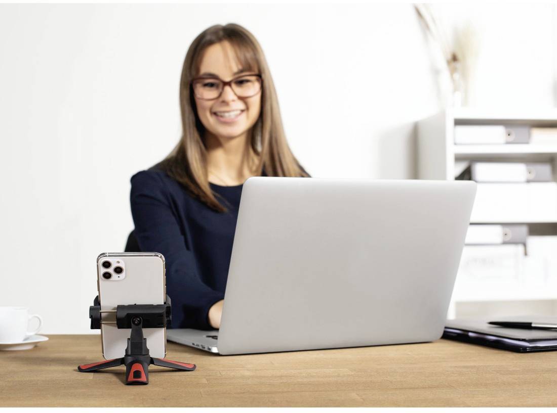 Eine Frau sitzt am Schreibtisch und arbeitet am Laptop. Vor ihr steht ein Smartphone auf einem Stativ. Im Hintergrund ist ein Regal.