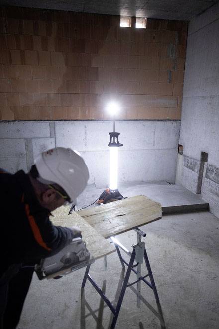 Eine Person in Schutzkleidung schneidet Holz mit einer Elektrosäge in einem halbfertigen Raum. Ein Bauleuchte beleuchtet den Raum.