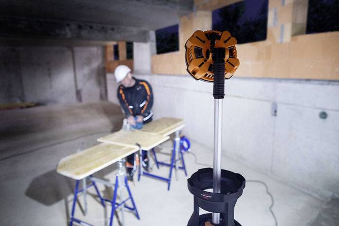 Ein Handwerker mit Schutzhelm arbeitet in einem Baustellenbereich an Holzstücken, wobei ein Baustrahler für Beleuchtung sorgt.
