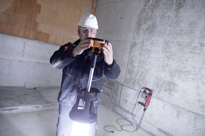 Ein Arbeiter mit Schutzhelm und Brille montiert eine Baustellenlampe in einem unvollendeten Raum mit Betonwänden.
