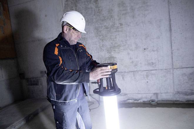 Ein Bauarbeiter trägt einen weißen Helm und bedient eine leuchtende Arbeitslampe in einem Betonraum.