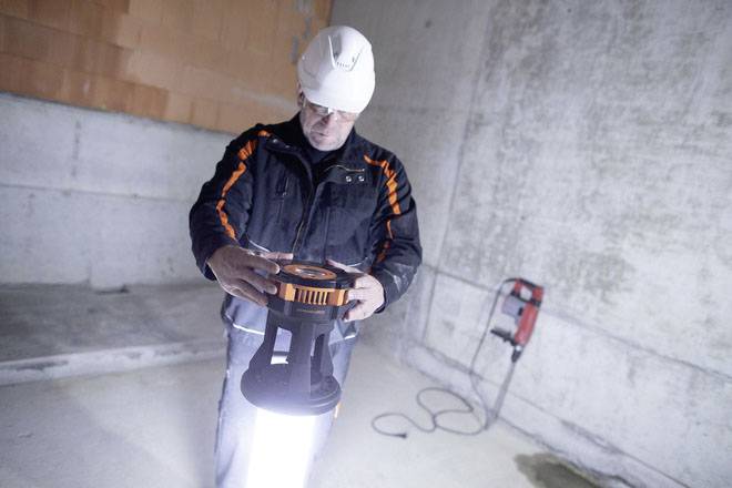 Ein Bauarbeiter in Schutzkleidung und Helm arbeitet in einem Betonkeller. Er hält eine leuchtende, runde Lampe in den Händen.