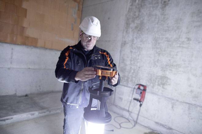 Ein Bauarbeiter in Schutzkleidung hält eine leuchtende Lampe in einem teilweise fertigen Raum mit Betonwänden.