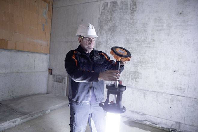 Ein Bauarbeiter in Sicherheitskleidung und Helm hält eine Arbeitsleuchte in einem unfertigen Betonraum hoch.