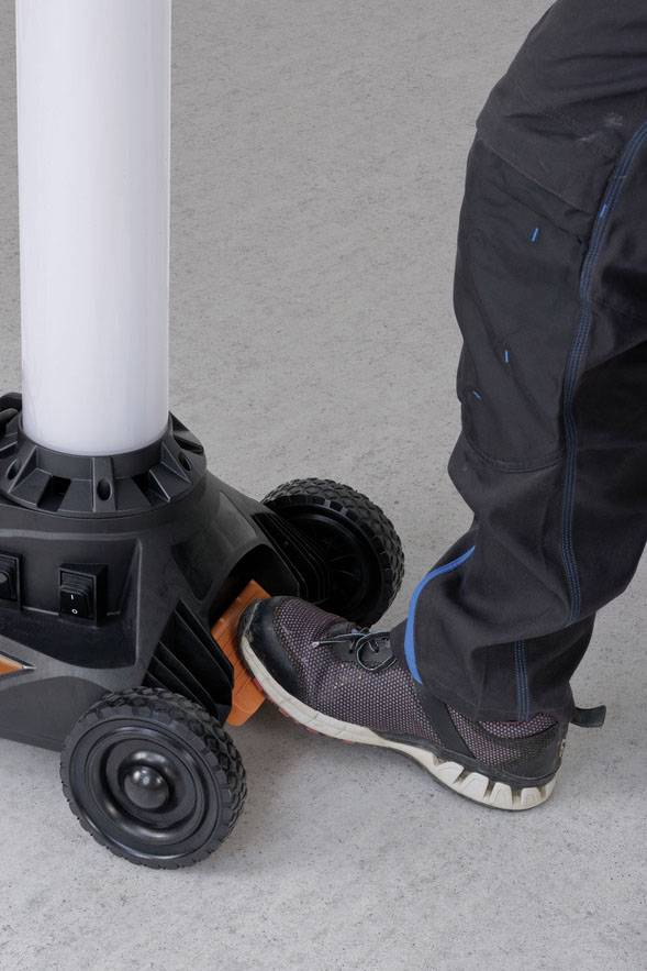 Person in dunkler Arbeitskleidung betätigt mit dem Fuß einen Schalter an einem Gerät mit Rädern, möglicherweise zur Stabilisierung.