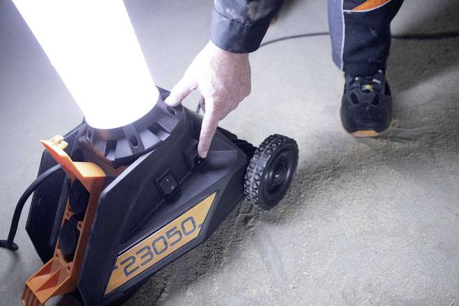 Eine Person schaltet eine Baustellenleuchte mit der Nummer '23050' ein. Die Lampe strahlt helles Licht aus und steht auf einem sandigen Boden.