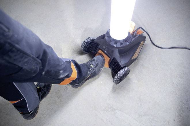 Person tritt mit einem Schuh gegen einen mobilen LED-Arbeitsstrahler auf Rädern. Der Fokus liegt auf der Interaktion mit dem Gerät.
