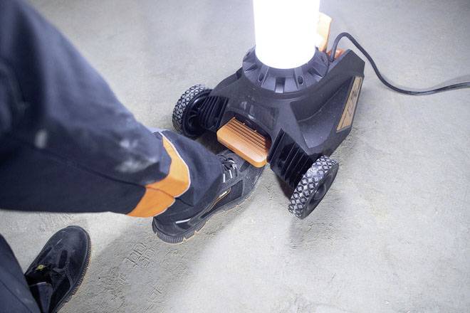 Eine Person tritt mit dem Fuß auf einen orangefarbenen Schalter an einem elektrischen Gerät, das auf einem Betonboden steht.