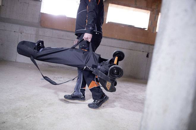 Eine Person trägt ein schwarzes elektrisches Skateboard in einer Tasche durch einen leeren Raum mit Betonwänden und Fenstern.