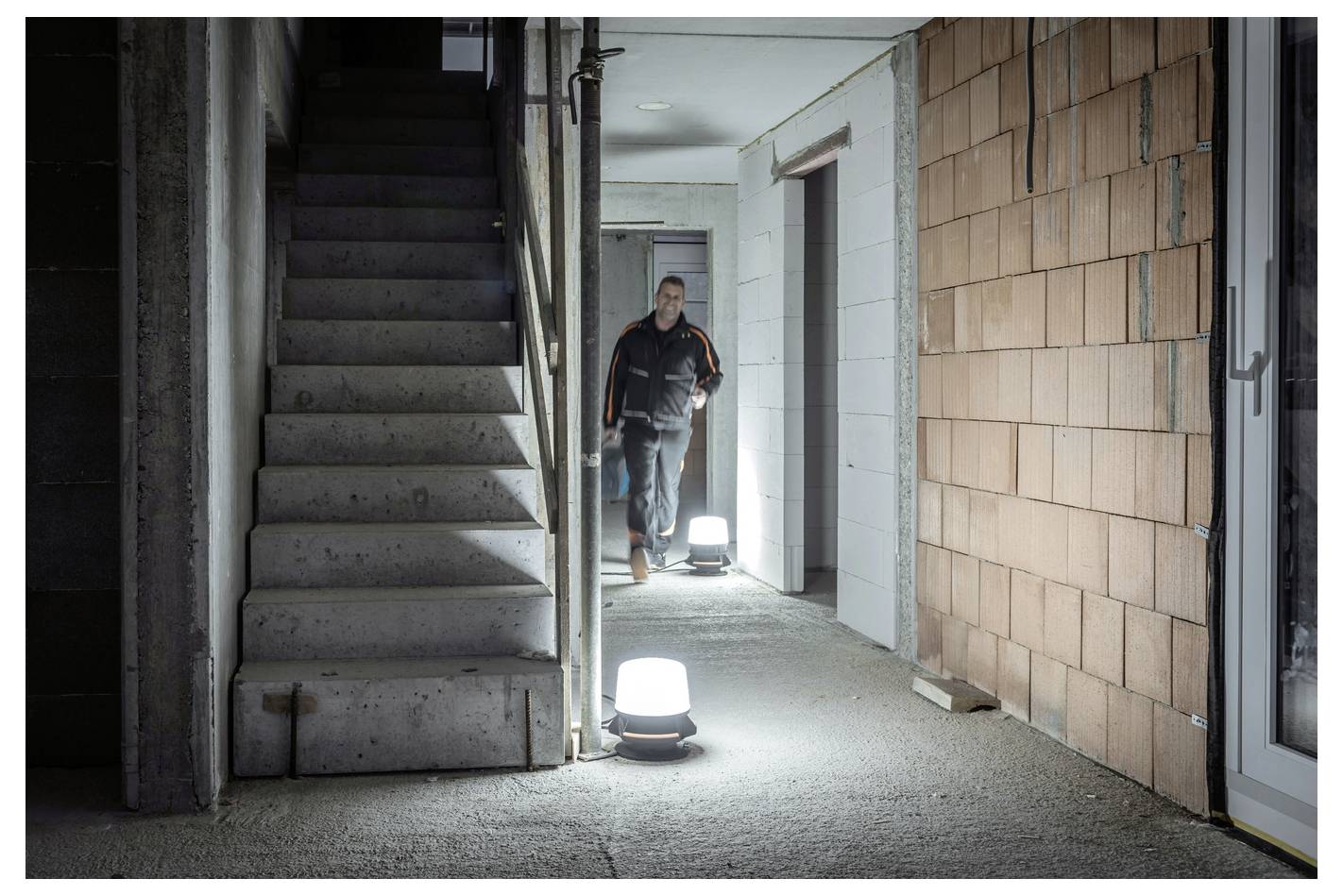 Ein Arbeiter steht in einem schwach beleuchteten unfertigen Flur neben Betontreppe und beleuchteten Stehlampen in einem Gebäude im Bau.