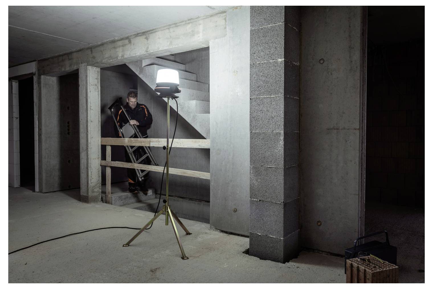 Bauarbeiter auf einer Leiter in einem unfertigen Betonkeller, beleuchtet durch ein Stativ-Arbeitslicht in der Nähe von Treppen.