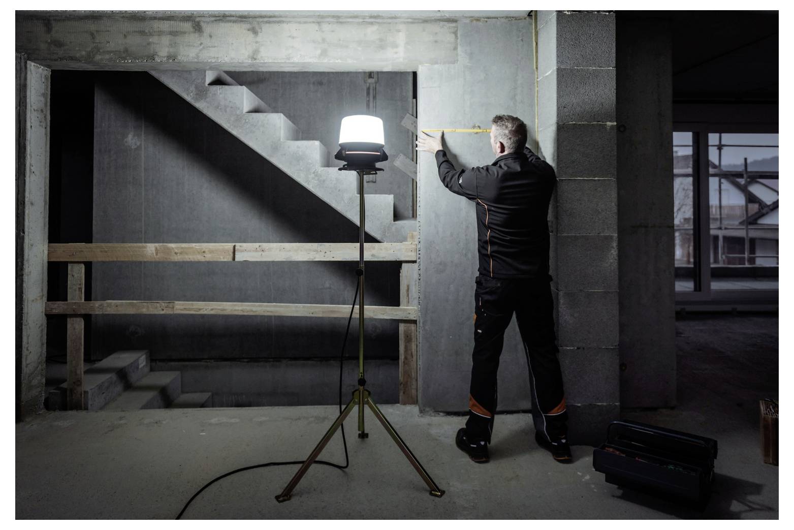 Ein Arbeiter misst eine Betonwand in einem unfertigen Gebäude, beleuchtet durch ein auf einem Stativ montiertes Baustellenlicht in der Nähe einer Treppe.