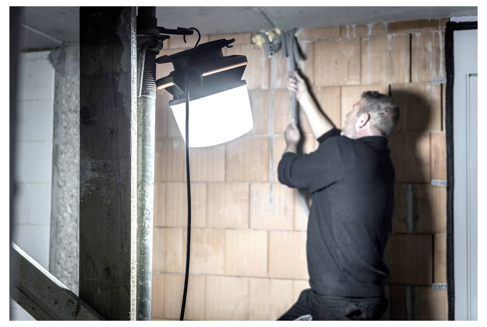 Ein Arbeiter installiert oder befestigt einen Draht an einer Backsteinmauer, während eine helle Arbeitsleuchte den Bereich beleuchtet.