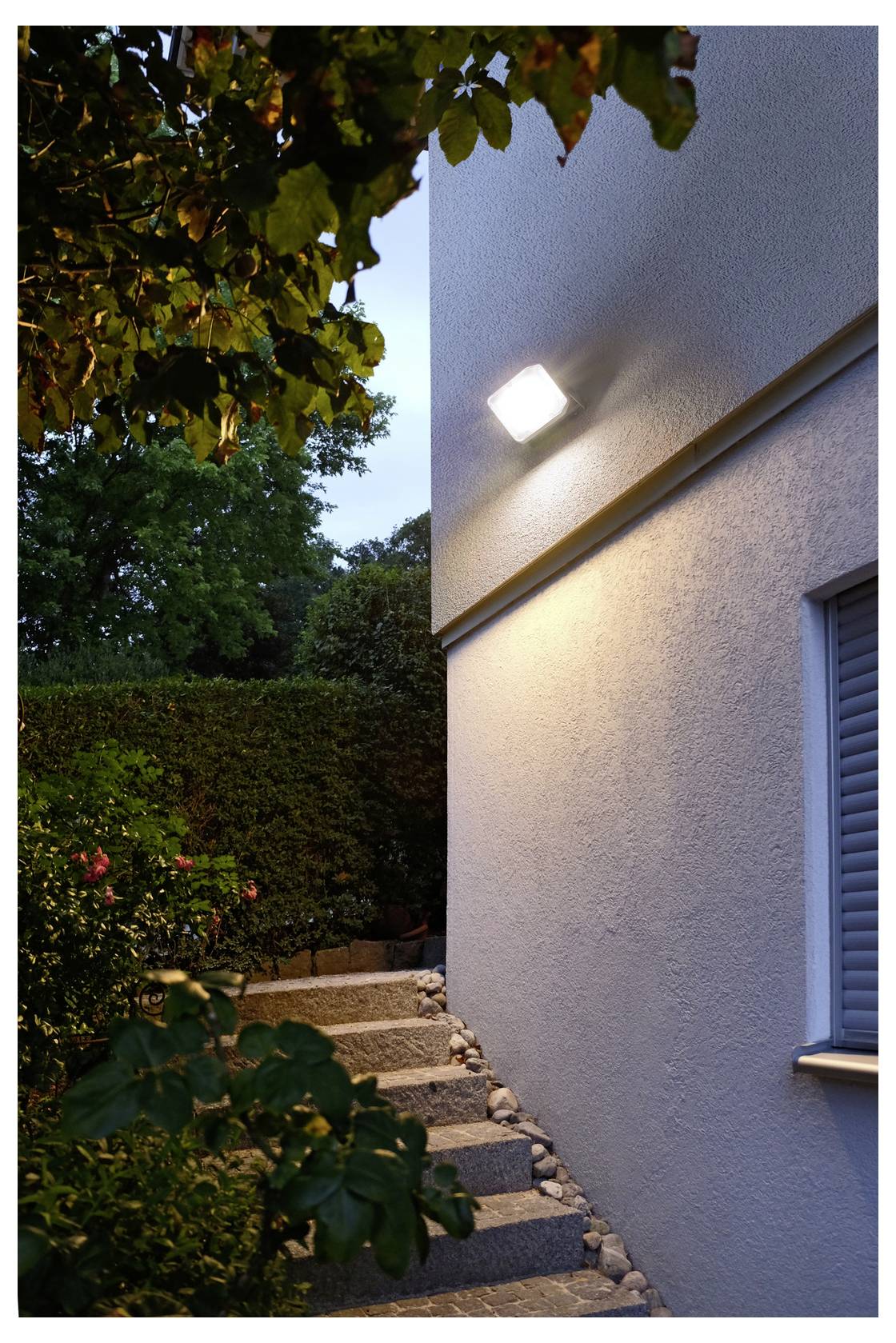 Außenseite eines Hauses in der Dämmerung, mit einer beleuchteten Wandleuchte auf einer Stuckwand neben einem schmalen Steinweg und Stufen durch einen blättrigen Garten.