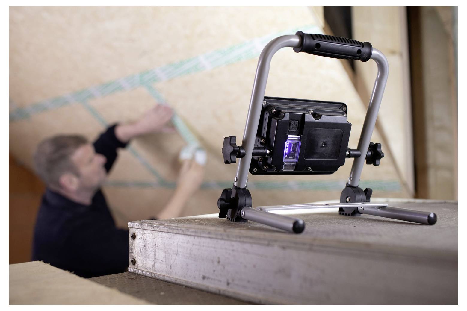 Eine tragbare Arbeitsleuchte auf einer Holzplattform im Vordergrund, mit einem unscharfen Mann im Hintergrund, der eine Wand misst oder markiert.