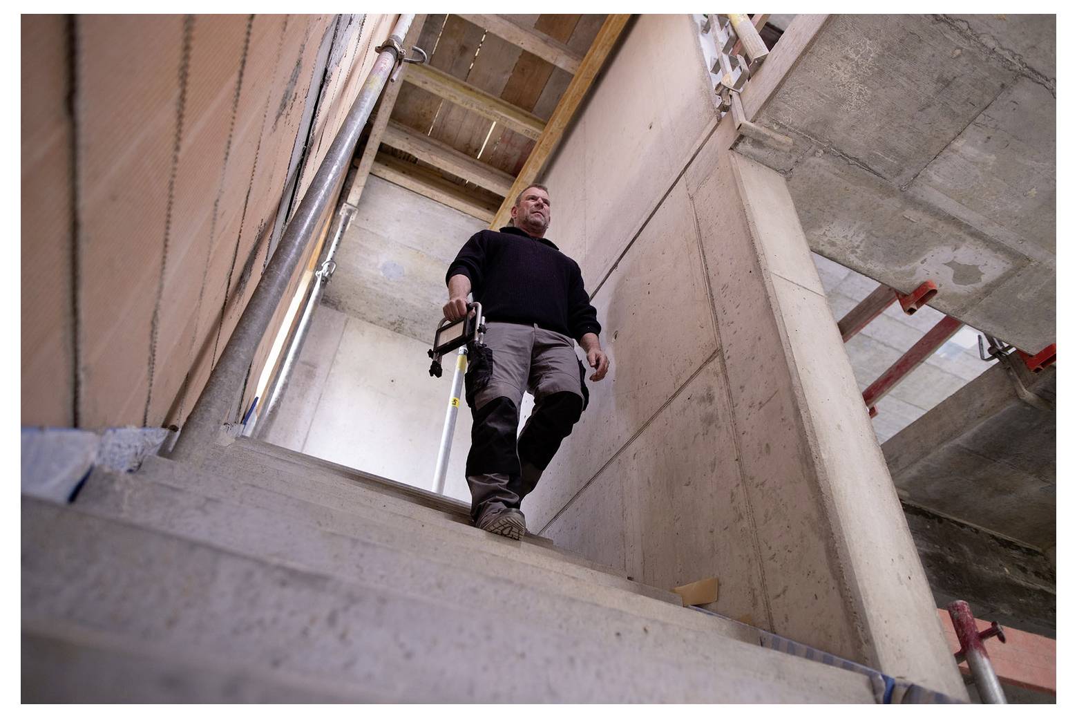 Mann, der Betontreppen auf einer Baustelle hinabsteigt, ein Werkzeug tragend, mit unfertigen Wänden und freiliegenden Strukturbalken um ihn herum.