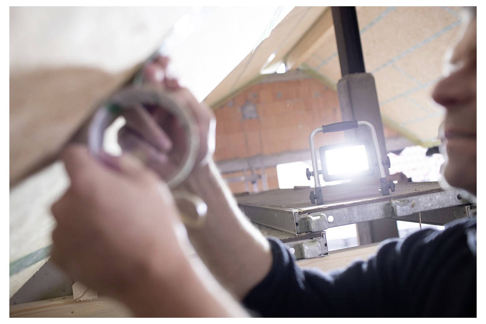 Eine Person hält ein kleines Objekt hochgehoben, um es in einer Werkstatt oder einem Zelt zu untersuchen, mit einer hellen Lampe und Ausrüstung im Hintergrund.