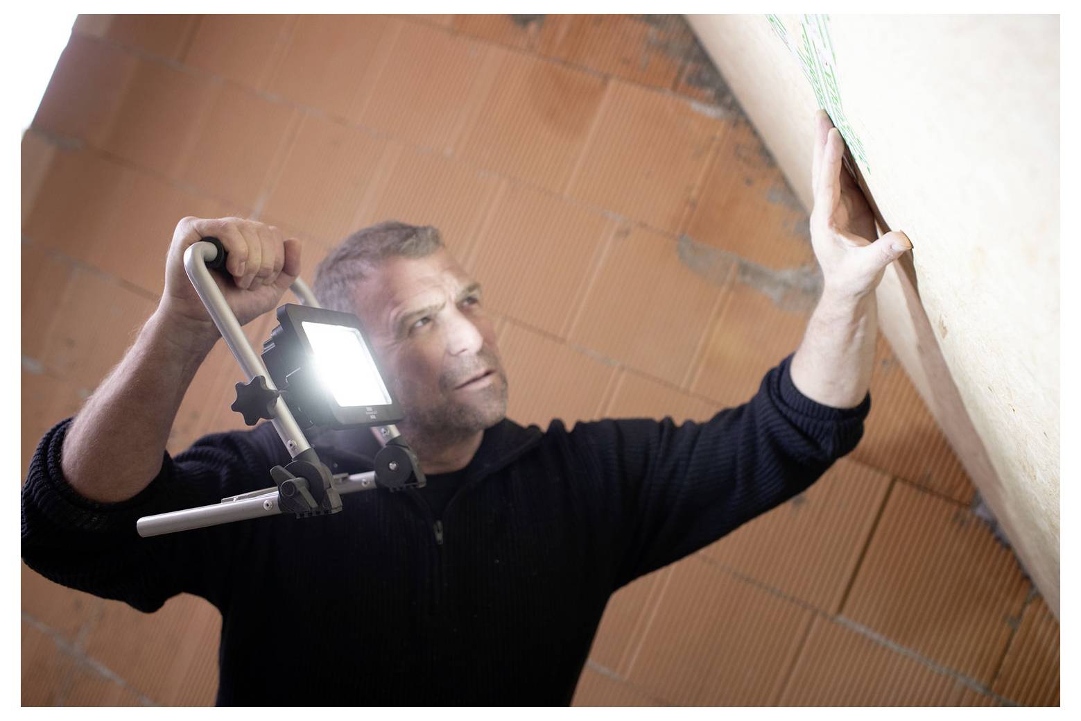 Ein Mann inspiziert eine Wand mit einer hellen Arbeitsleuchte, während er die Lampe hält und sich nach oben streckt, wahrscheinlich während einer Hausreparatur oder Installation.