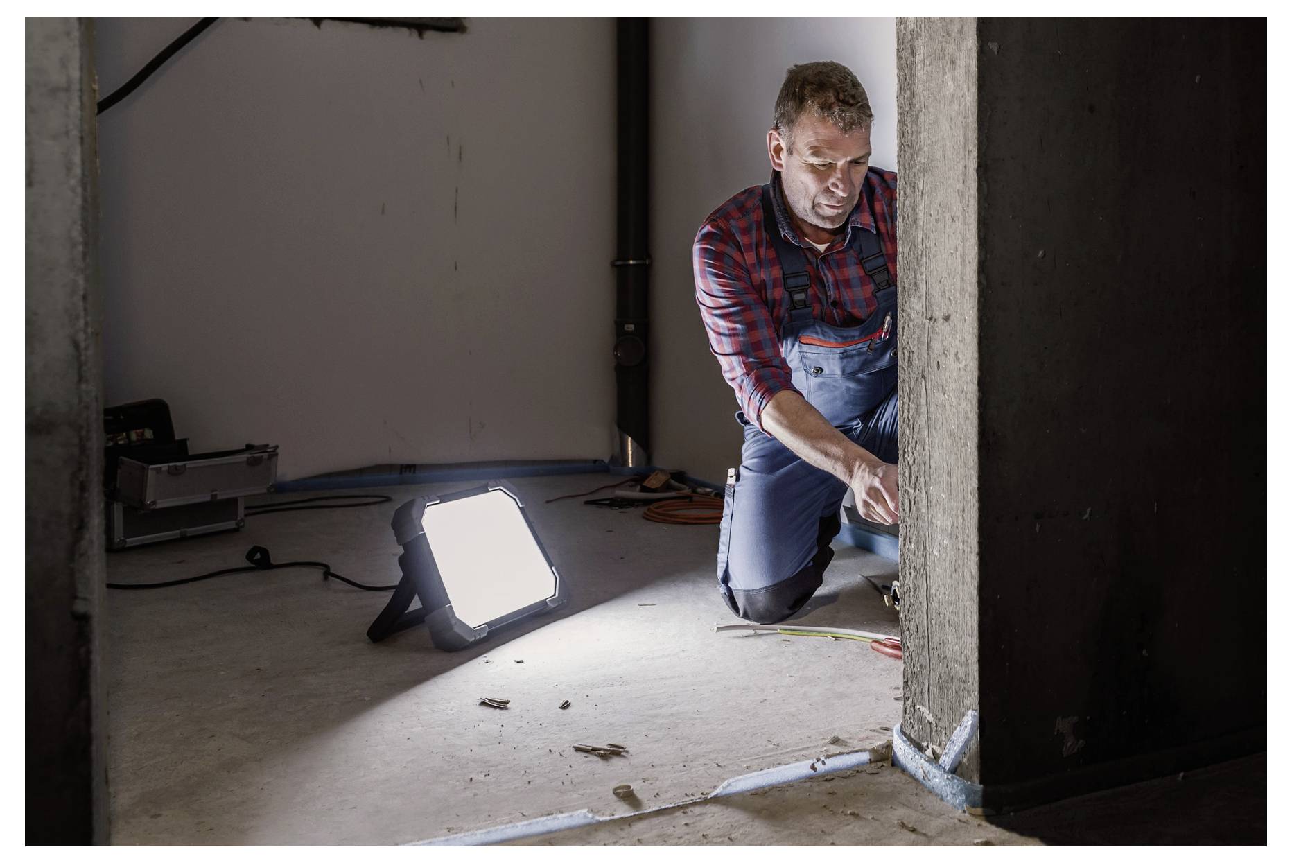 Ein Mann kniet neben einer Wand und nutzt Werkzeuge, um an Stromleitungen oder Reparaturen zu arbeiten, beleuchtet durch ein helles tragbares Arbeitslicht in einem unvollendeten Raum.