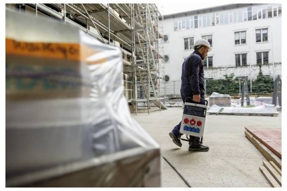 Bauarbeiter trägt eine Werkzeugkiste über einen mit Gerüsten gesäumten Hof auf einer Baustelle.