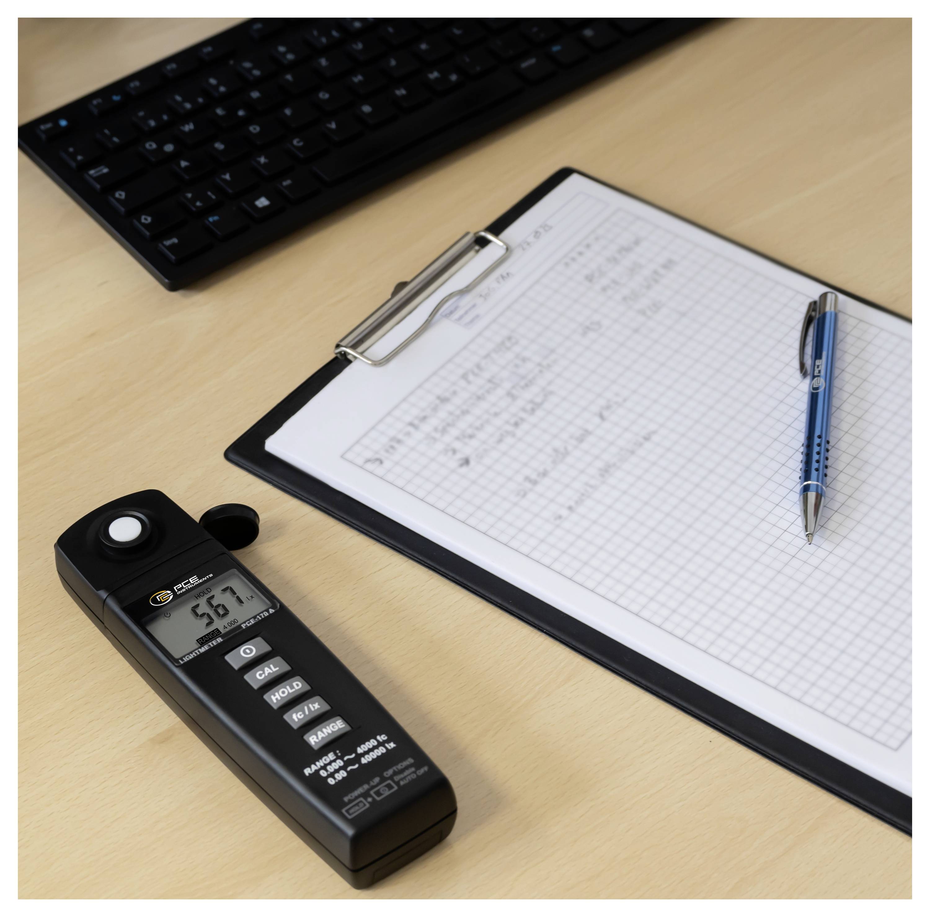 Luxmetermessung 56,7 auf einem Schreibtisch neben einer Zwischenablage mit Millimeterpapier, einem Stift und einer Tastatur.