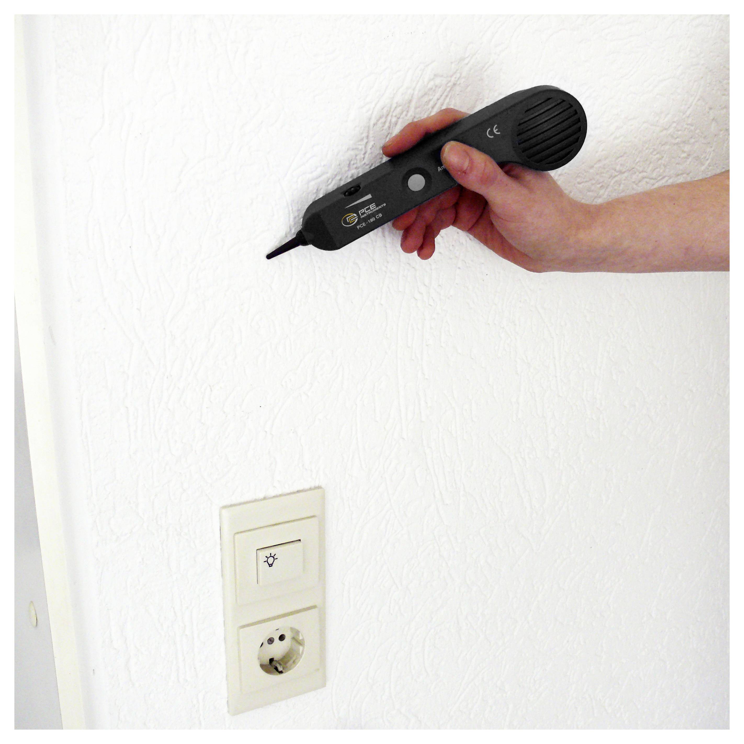 Eine Hand hält ein Spannungsprüfgerät in der Nähe einer Wand über einem Lichtschalter und einer Steckdose und prüft die Wand auf stromführende Leitungen.