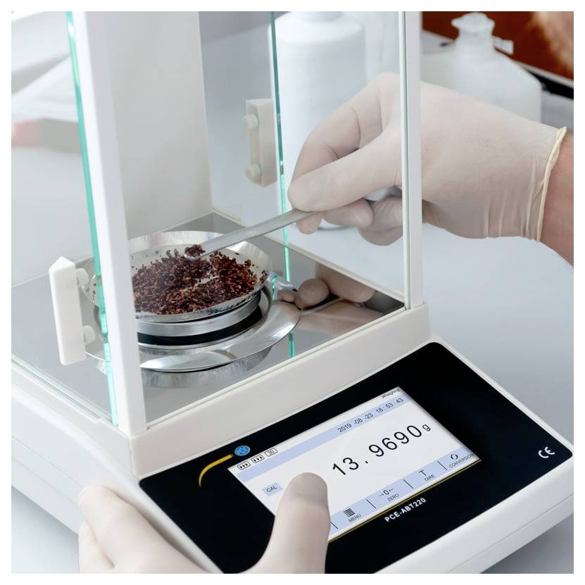 Behandschuhte Hände bedienen eine digitale Präzisionswaage und wiegen eine kleine Probe in einem Laborinstrument.
